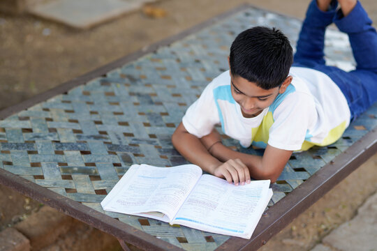 Indian schoolboy lying on a traditional charpai while studying from a textbook outdoors in a peaceful rural setting.