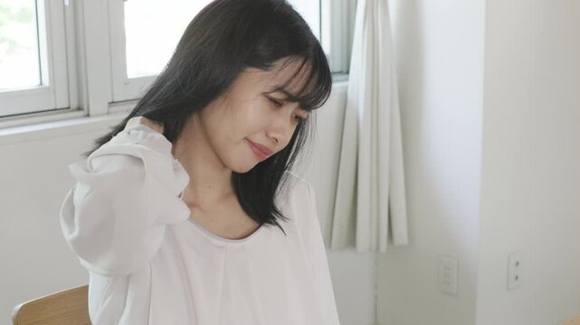 Asian woman frowning while touching her shoulder in bright room