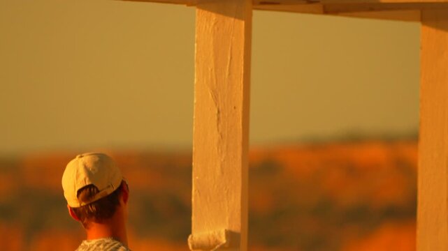 Painting renovation sunset man applies white paint to wooden pillars of a coastal structure during a bright orange golden hour