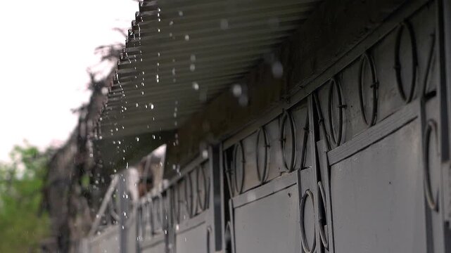 Close-up of raindrops falling from the roof edge beside a gray exterior wall, with circular window grilles visible in soft overcast light and a damp urban