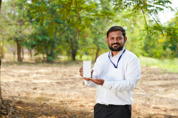 Fototapeta premium Indian businessman standing in a rural field holding a smartphone with a blank screen and showing a thumbs up gesture.