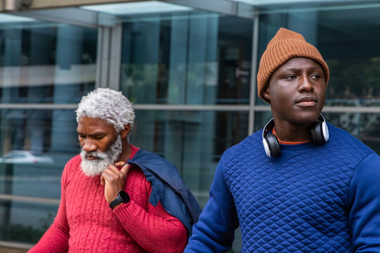 African American men walking sidewalk past glass facade, wearing beanie and headphones, copy space
