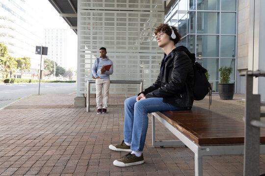 Diverse teenage male students waiting at shelter bench with headset, holding red folder, copy space