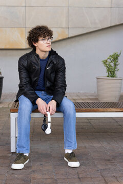 Man sitting on wooden bench in courtyard wearing leather jacket holding headphones with glasses