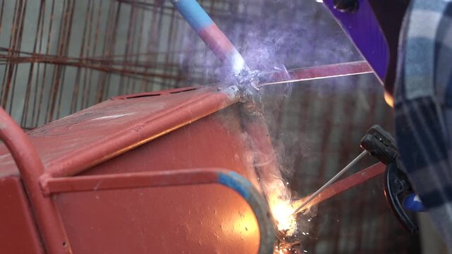 Close-up of a worker using a welding torch to join metal parts on a red painted tank or barrel, with intense sparks, glowing heat, and a blurred factory