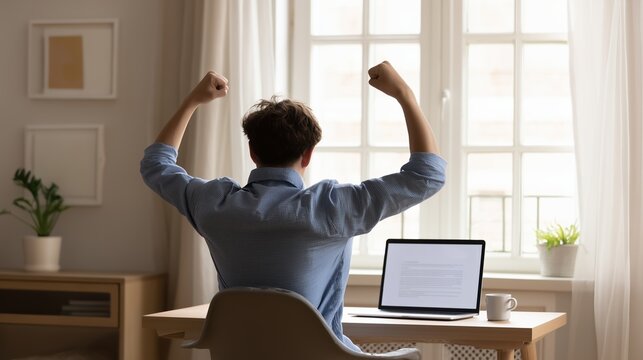 A man sitting at a home desk raises both fists in triumph while facing an open laptop, expressing success and excitement in a bright, modern workspace setting.