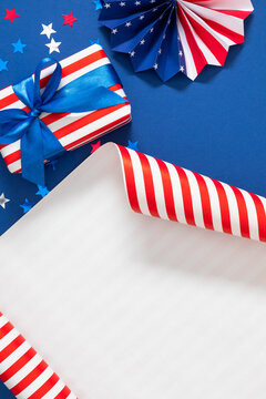 Fourth of July gift box with wrapping paper roll and patriotic paper fan on blue background with star confetti. 4th of July Independence Day vertical flat lay backdrop with large white copy space.