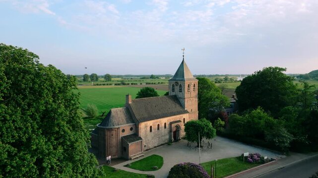 Aerial view of the historic Oude Kerk in Oosterbeek, Netherlands, showcasing its ancient architecture amidst lush green fields and trees under a serene blue sky. Captures the tranquil village setti...