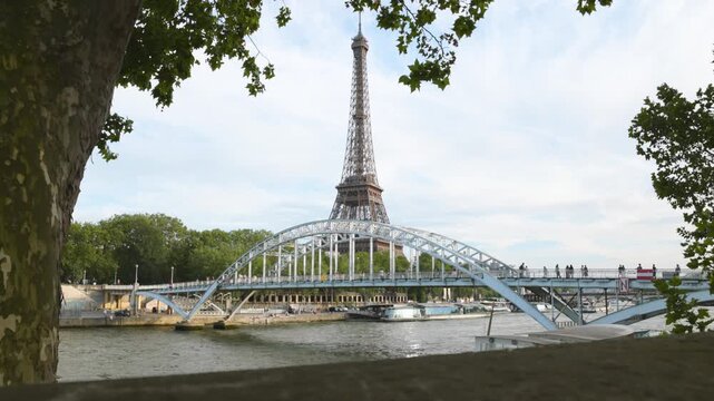 A picturesque view of the Eiffel Tower perfectly framed by a stone wall, green leaves, and the elegant arched Debilly Footbridge over the Seine River.