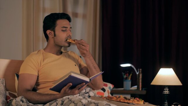 Modern well-groomed boy eating pizza while reading an enjoyable storybook. Medium shot of an urban Indian male studying in the light of a night lamp relaxing in his bedroom
