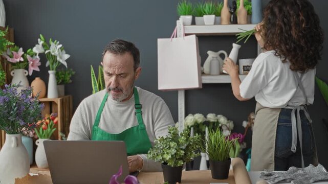 Man florist in green apron typing on laptop while woman arranges tulips and holds a vase in a flower shop; focused teamwork.