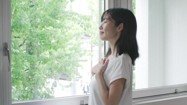 Young Asian woman placing hand on chest while breathing deeply by window