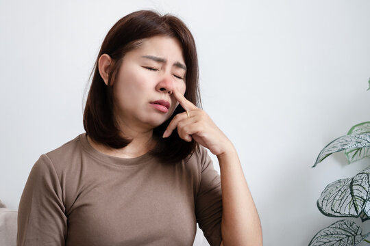 Asian woman touching her nose with a painful expression, suffering from sinus infection or allergies. Concept of nasal congestion, cold and flu, allergic rhinitis, and respiratory illness
