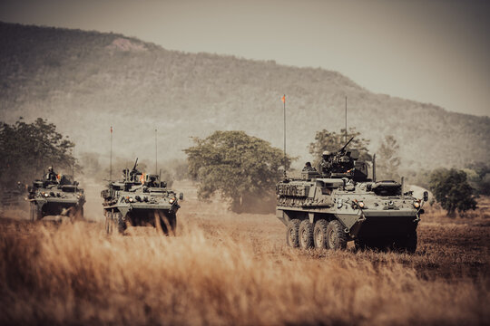 Dramatic shot of an eight-wheeled green military armored vehicle (APC) advancing through a dense dust and haze cloud on a dry dirt field under heavy golden sunset light.