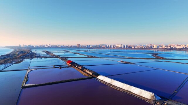 Aerial view of salt evaporation ponds near Burgas, Bulgaria. Elevated perspective of geometric blue and purple pools, white salt stockpile, red-roofed building, city and sea on horizon.
