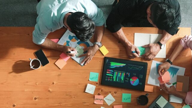 Top aerial view of diverse team writing colorful sticky notes to brainstorm marketing idea while looking at financial chart. Multicultural smart business people taking a note about plan. Convocation.