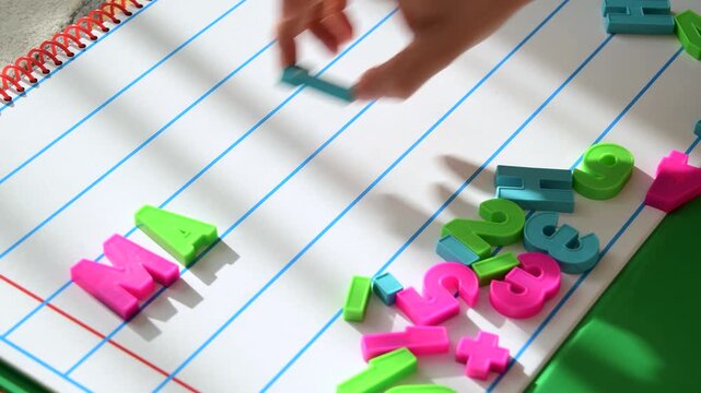 Child's hand spelling the word MATH with colorful plastic letters on lined notebook paper. Education, literacy, and numeracy learning concept