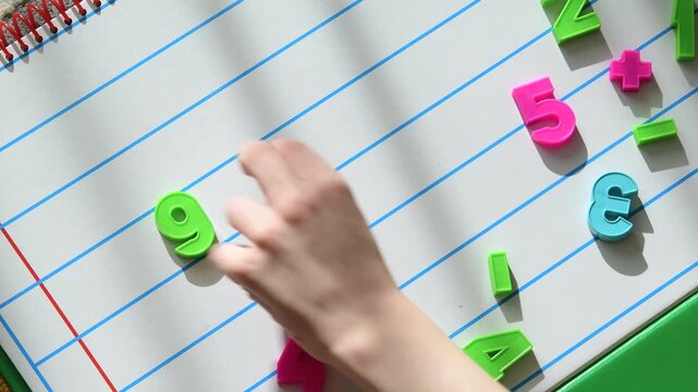 Child's hand arranging colorful plastic numbers and math symbols on lined notebook paper, early learning and education concept