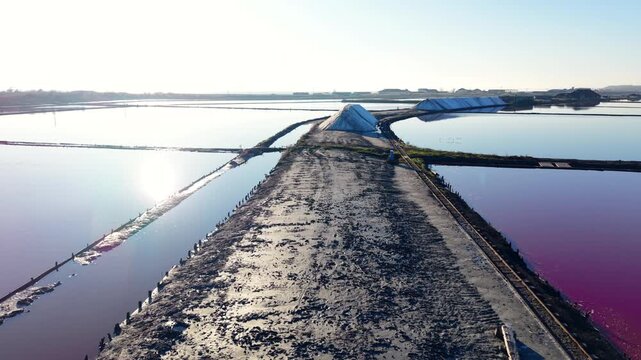 Drone view of salt farm embankment between colored ponds, Bulgaria. Elevated perspective along a central dirt levee flanked by blue and magenta pools, salt pile at midpoint, flat horizon.