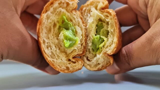 Sidoarjo, Indonesia - 4 June, 2026: Hands breaking apart a freshly baked flaky croissant filled with sweet green cream for a delicious morning snack.