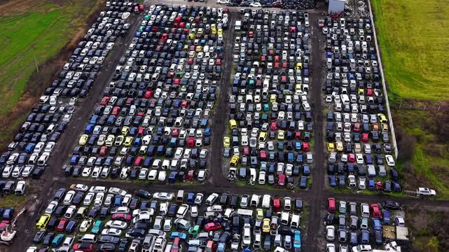 Wide aerial view of large vehicle salvage yard, Bulgaria. High elevated drone perspective over hundreds of colorful wrecked cars in multiple rows with green fields on all sides.