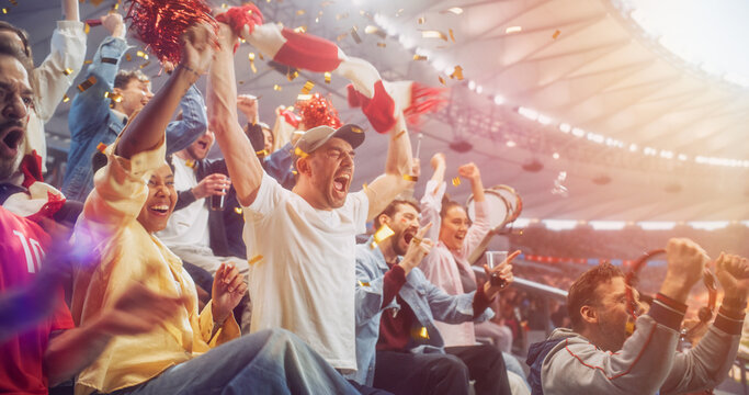 Diverse Soccer Supporters Surge Upright in Stadium Seats During a Football Championship, Shouting in Disbelief, Relief and Victory While a Red Scarf Swings, a Tambourine Shakes and Confetti Falls.