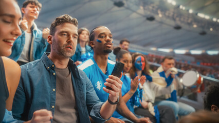 Obraz premium Portrait of a White Male Fan Checking a Smartphone in Stadium Stands at a Soccer Football Championship Tournament, Surrounded by a Happy Excited Diverse Crowd of Smiling Supporters Cheering a Goal.