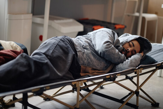 Young adult man resting on camp bed in humanitarian aid center, receiving temporary shelter support during crisis displacement. Useful for refugee relief, emergency housing campaigns