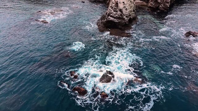 High angle perspective of white foam and churning turquoise water around stone boulders. Ocean waves crashing against large rocks in the sea.