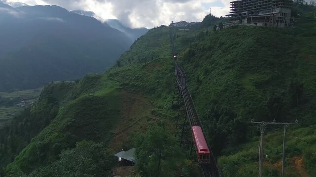 The Sapa red mountain train exits a tunnel through a mountain as another train approaches from the other direction, aerial wide view on a cloudy day around dusk