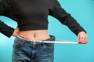 Confident woman holds tape measure around slim waist showing healthy weight loss journey with oversized jeans against blue background