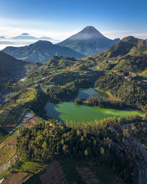 Aerial view of the vibrant green Telaga Warna lake surrounded by lush forests and terraced agricultural fields with majestic volcanoes under a clear blue sky in Indonesia.