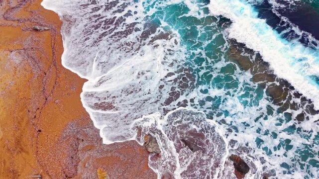 Straight down drone view of white ocean foam breaking on a wide empty coastal shoreline. Overhead perspective of waves washing onto reddish sandy beach.