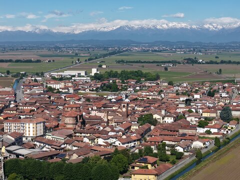 Aerial view of the historic town center with red-tiled rooftops and a church dome against the backdrop of snow-capped Alps in San Germano Vercellese, Piedmont, Italy.