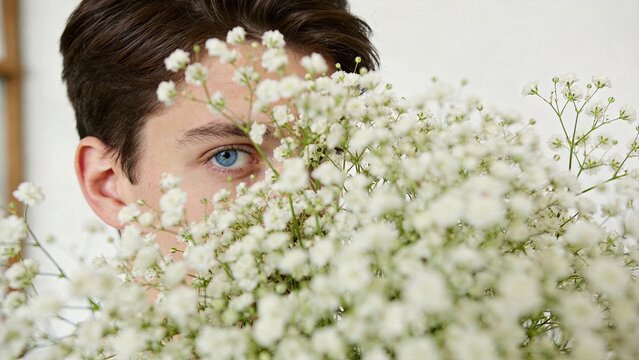 Close-up portrait of a young man with striking blue eye peeking through a bouquet of delicate white baby's breath flowers. Natural beauty, spring freshness, romantic gesture,