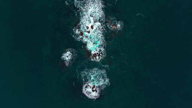 Aerial view of white surf swirling around submerged dark stones in dark teal ocean waters. Overhead perspective of isolated reef rocks in deep sea.
