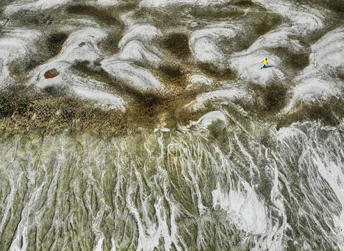 Aerial view of a person in a yellow shirt walking across a textured landscape of dried mud and white mineral deposits in Bogura, Bangladesh.