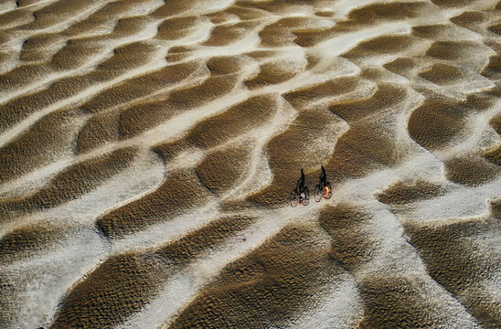 Aerial view of two people riding bicycles across a vast textured sandy landscape with intricate ripple patterns and long shadows in Bogura, Bangladesh.