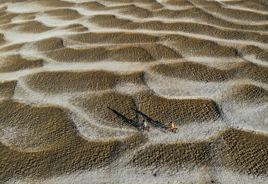 Aerial view of two people riding bicycles across a vast textured landscape of sand dunes and dried mud patterns with long shadows in Bogura, Bangladesh.