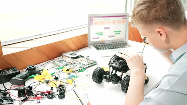 Happy teenager fixing robotic model while using electronic equipment remove car wheel with wire and laptop scatter around. Smart boy study or learn about car robot construction at class. Edification