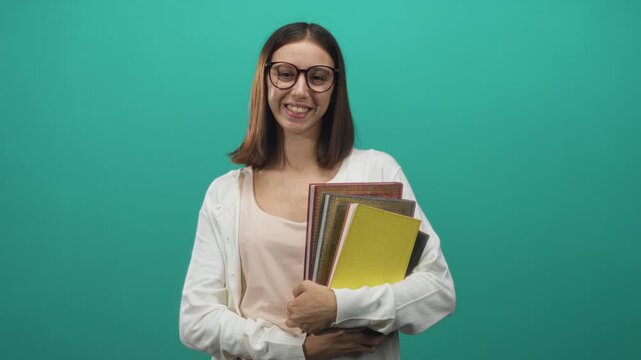 Woman smiling while holding a stack of colorful notebooks and textbooks, hands clasped across chest wearing round glasses in studio; cheerful focused learning.