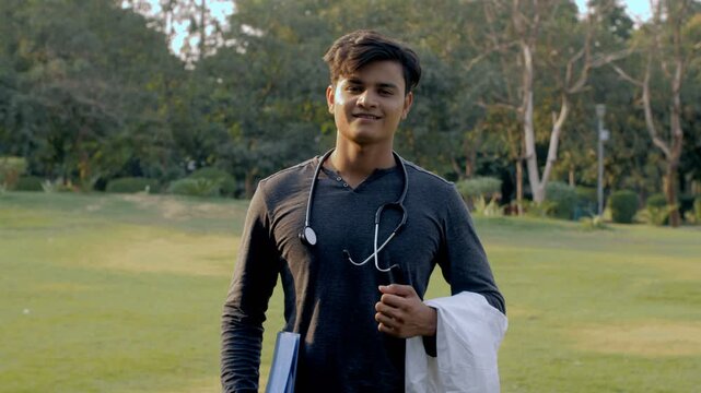 Young aspiring male doctor with a stethoscope on the neck standing in the park. Medium shot of a charming studious boy with a stethoscope  a doctor's overcoat  and a book in an open area
