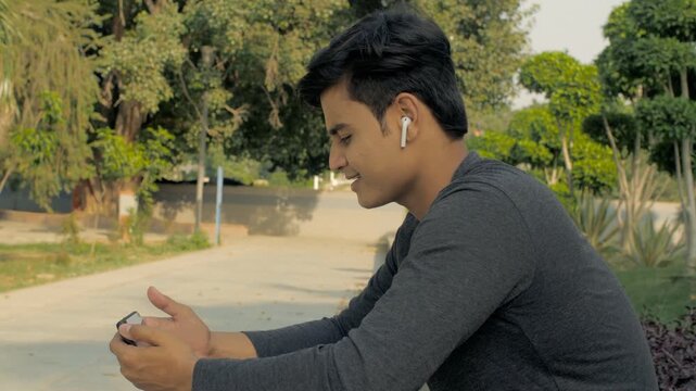Lively Indian man listening to his favorite music while relaxing inside a park. Closeup shot of an energetic young male enjoys hearing peppy songs using a bluetooth earphone - relaxation concept