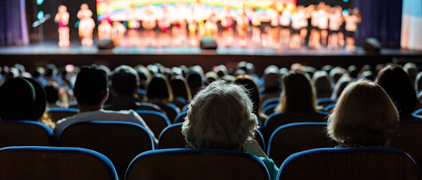 Audience sitting in theater rows watching performance on stage. People enjoying concert in auditorium. View from back of crowd