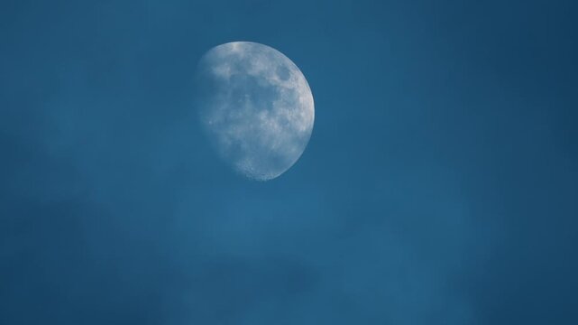 Cinematic close up video of the daytime moon shining through thin clouds in the blue sky.
