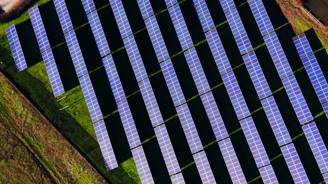 Elevated perspective of renewable electricity generators casting dark linear shadows on green pasture. Geometric pattern of parallel solar panel rows on grass.