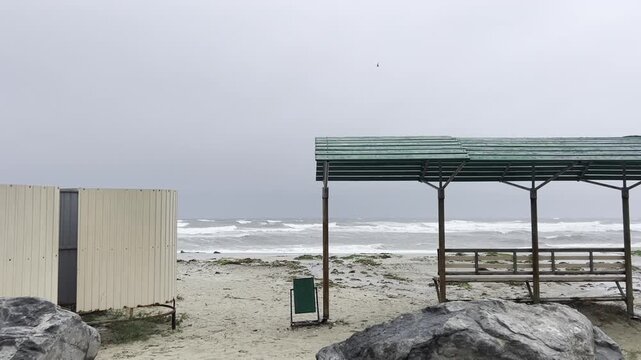 Stormy sea with rough waves crashing on a deserted sandy beach. Gloomy weather over the Pacific coastline with a wooden shelter and changing cabin near ocean.