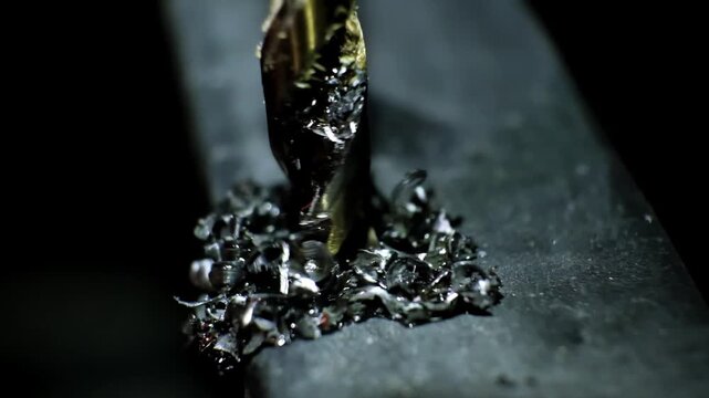 Close-up macro shot of a metallic twisting drill bit drilling a dark metallic surface, stained with metal shavings and lubricant. Metal processing. Construction works. B-roll 4K slow motion footage