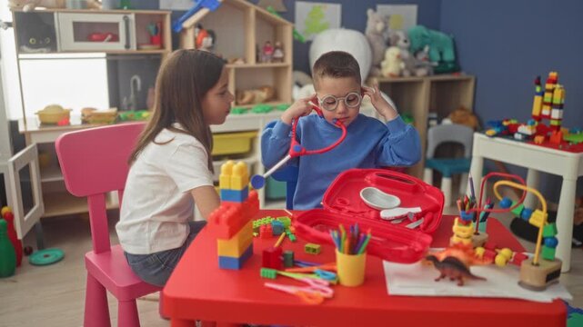 Girl reaches to hand a toy stethoscope to a boy wearing glasses while seated at a red table with a toy doctor kit in a studio playroom; curiosity learning cooperation.