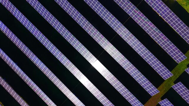 High angle pattern composition of electrical modules over dark ground with bright sunbeams striking the surface. Diagonal view of sustainable solar farm rows from above.